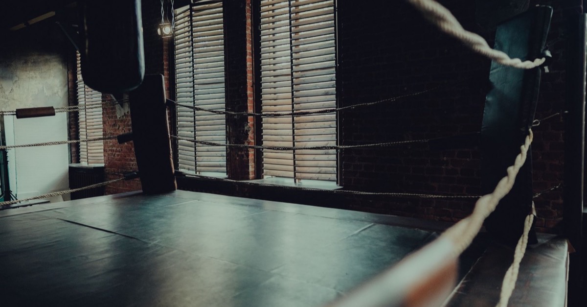 Corner of an empty boxing ring in a dim gym with brick walls and window blinds