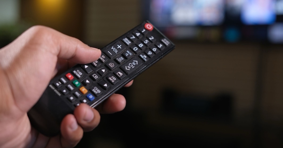 Hand pointing a black TV remote at a blurred television screen